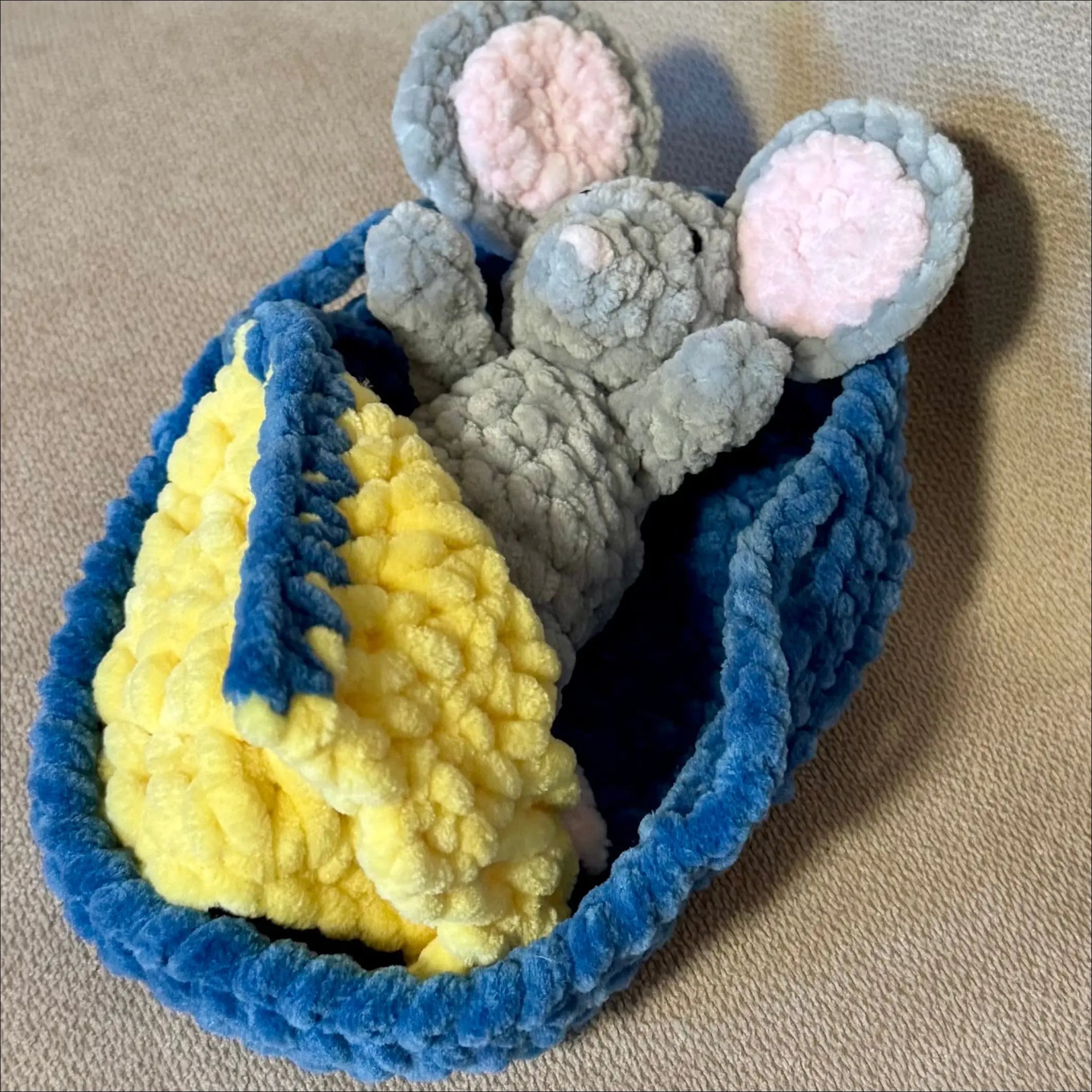 A handmade crocheted baby blanket set featuring a blue and yellow blanket wrapped around a gray plush mouse toy with pink inner ears, displayed on a beige fabric background.