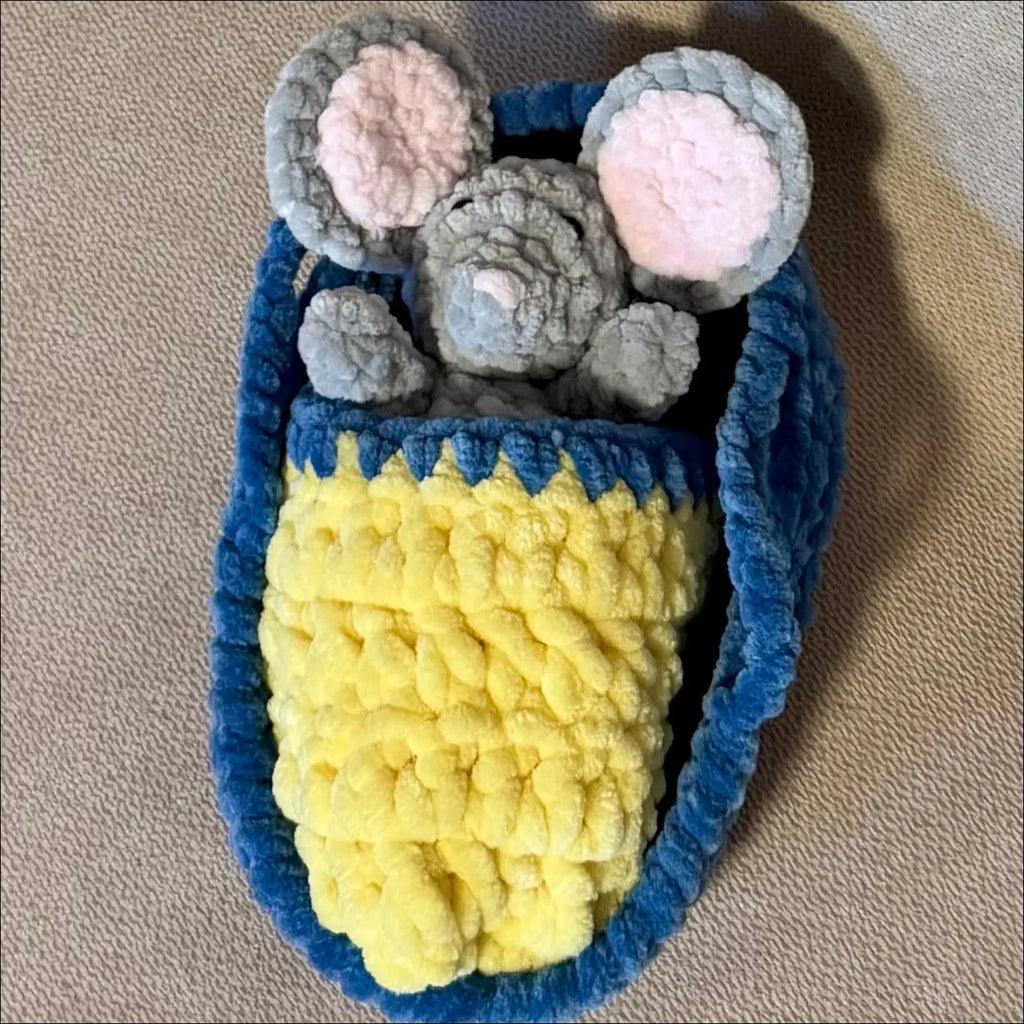 A handmade crocheted or knit animal-themed pouch or bag featuring a gray mouse head with pink inner ears on top of a yellow body and blue border, placed on a beige textured fabric background.