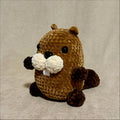 A small crocheted beaver toy sits centered on a beige fabric background. The beaver is brown with cream-colored tusks and black bead eyes.