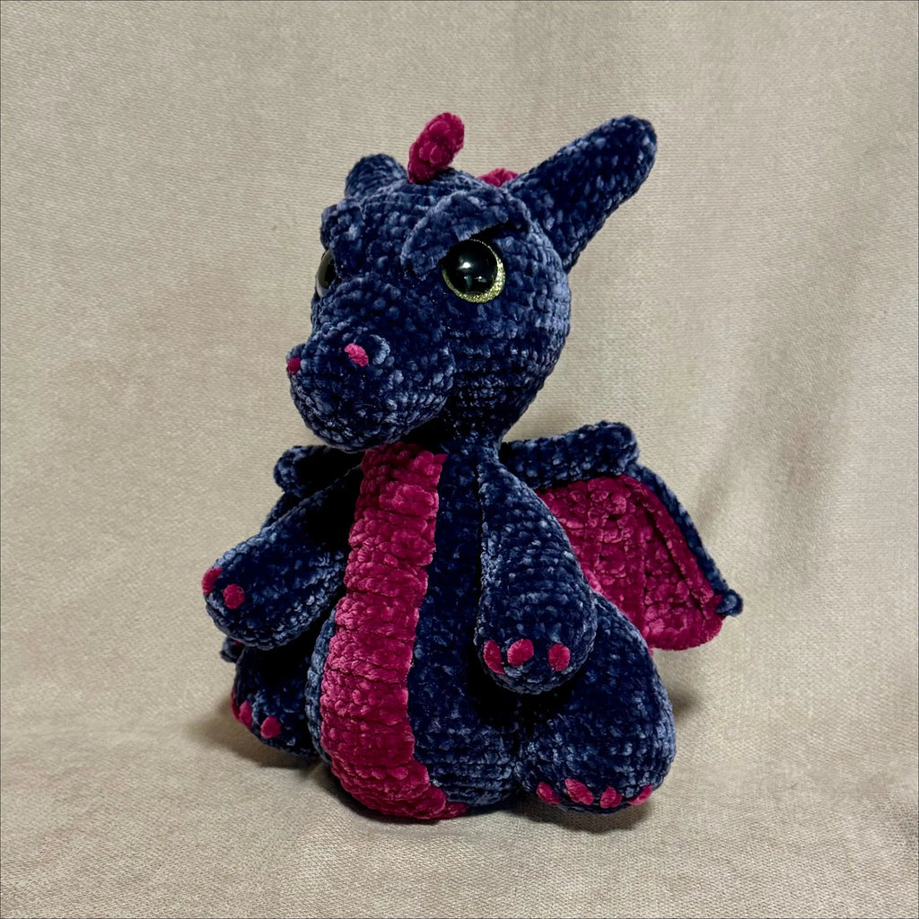 A handmade crocheted dragon toy in dark blue and magenta pink, sitting against a neutral beige fabric backdrop.