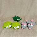Three small crocheted plush toys on a beige fabric background: two green turtle-like characters and one dark green crocodile-like character, plus two gray mouse-like characters with pink ears and pink noses. No visible brand markings.