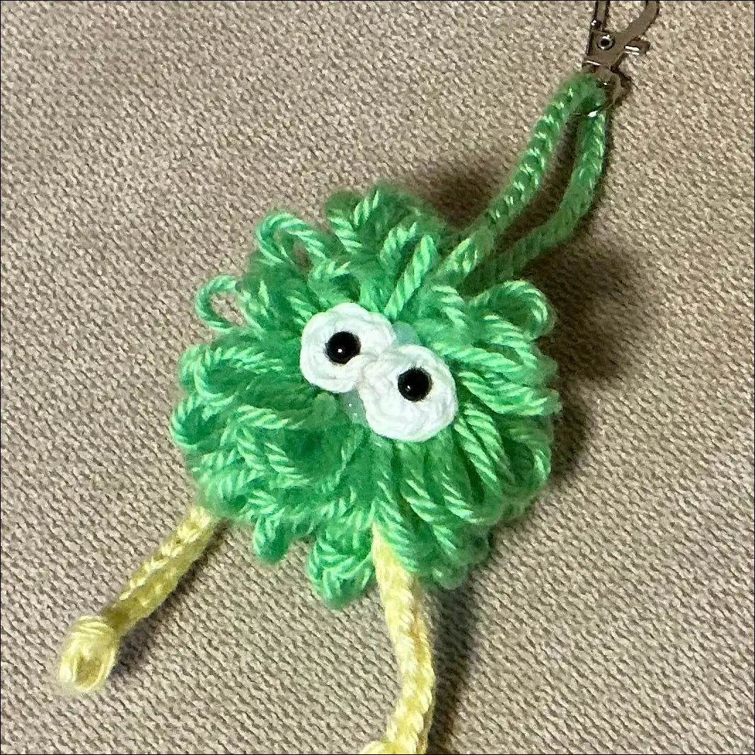 A handmade green yarn monster keychain with googly eyes and yellow legs, resting on a textured beige fabric background.