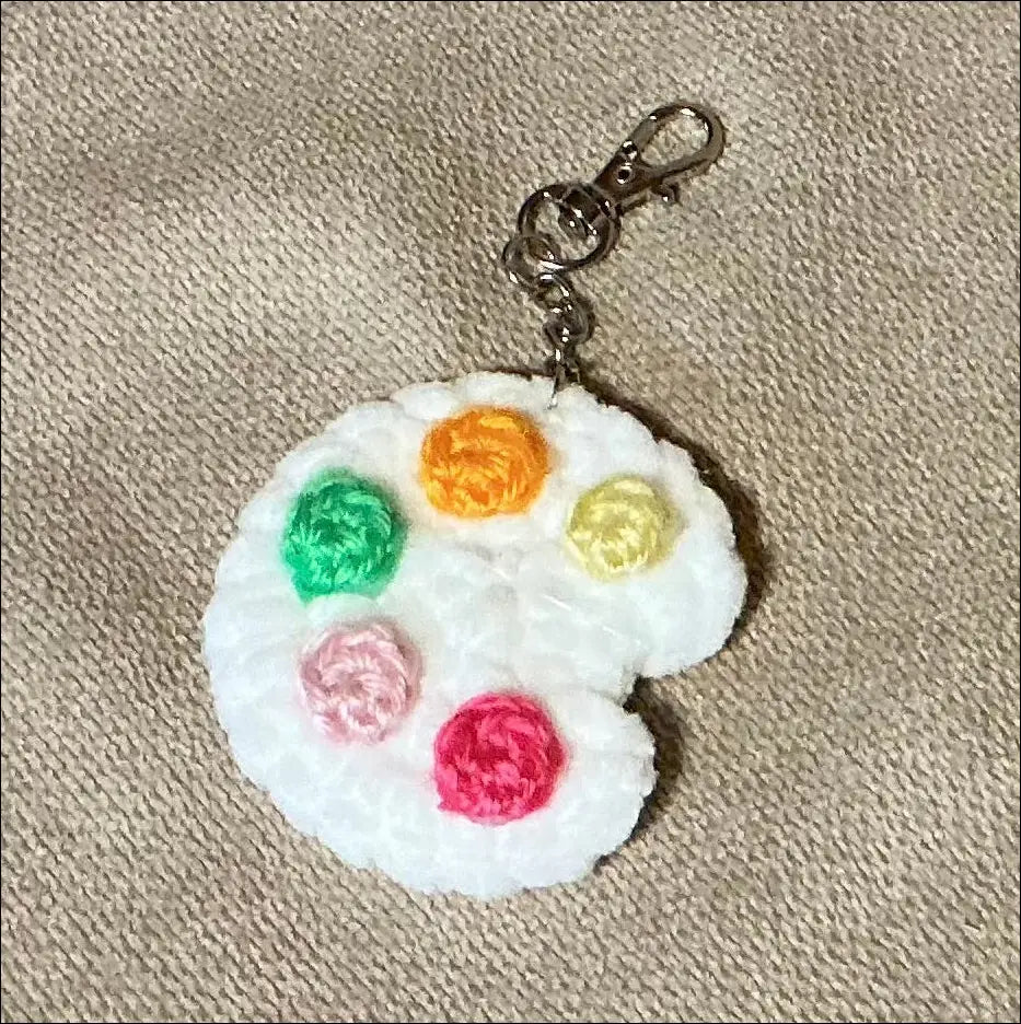 A crocheted white paint palette charm with five colorful painted color circles in green, orange, yellow, pink, and red, attached to a silver metal lobster clasp, displayed on a textured beige fabric background.