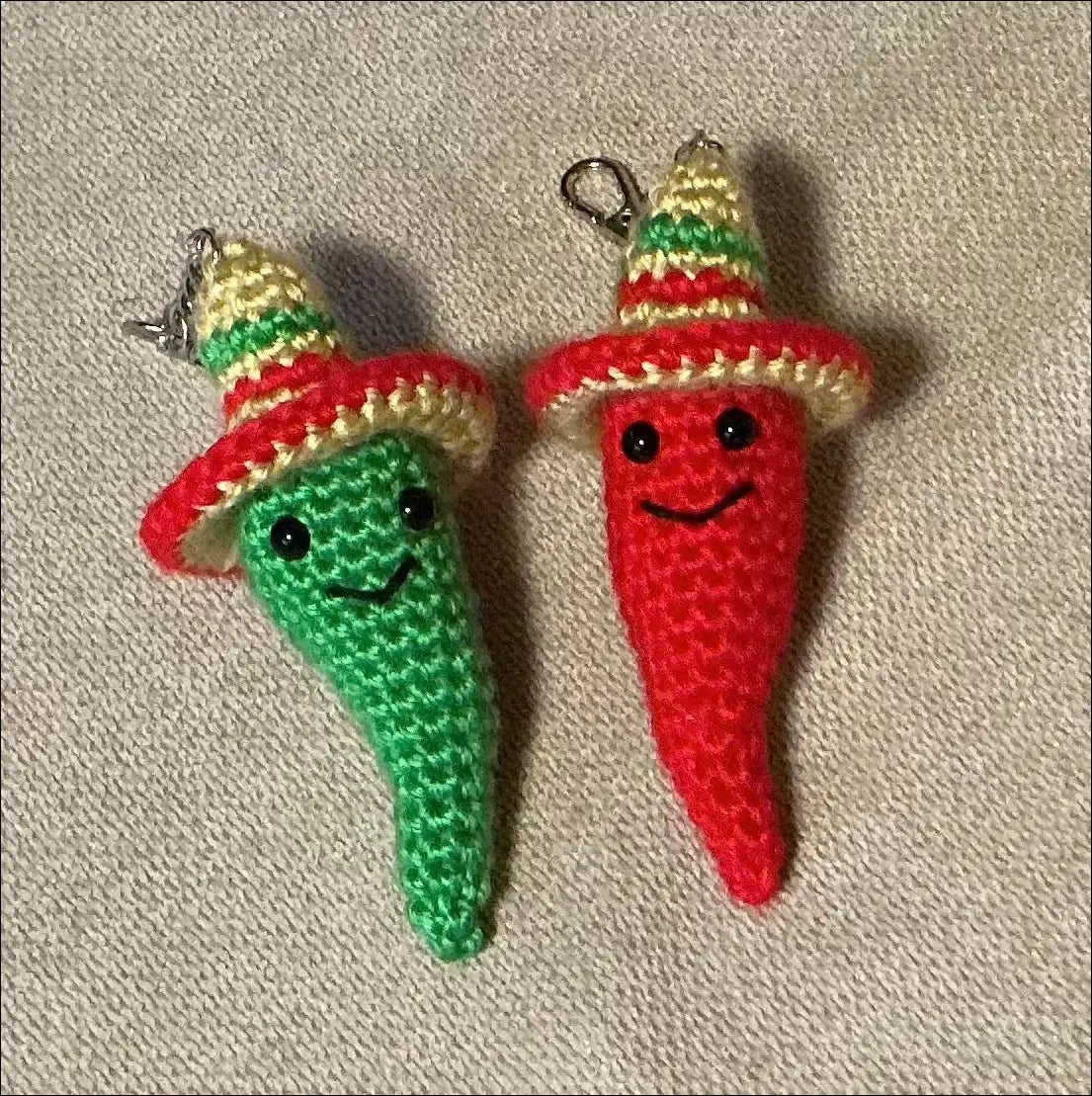 Two small crocheted chili pepper charms with smiling faces and colorful sombreros, placed on a beige fabric background.