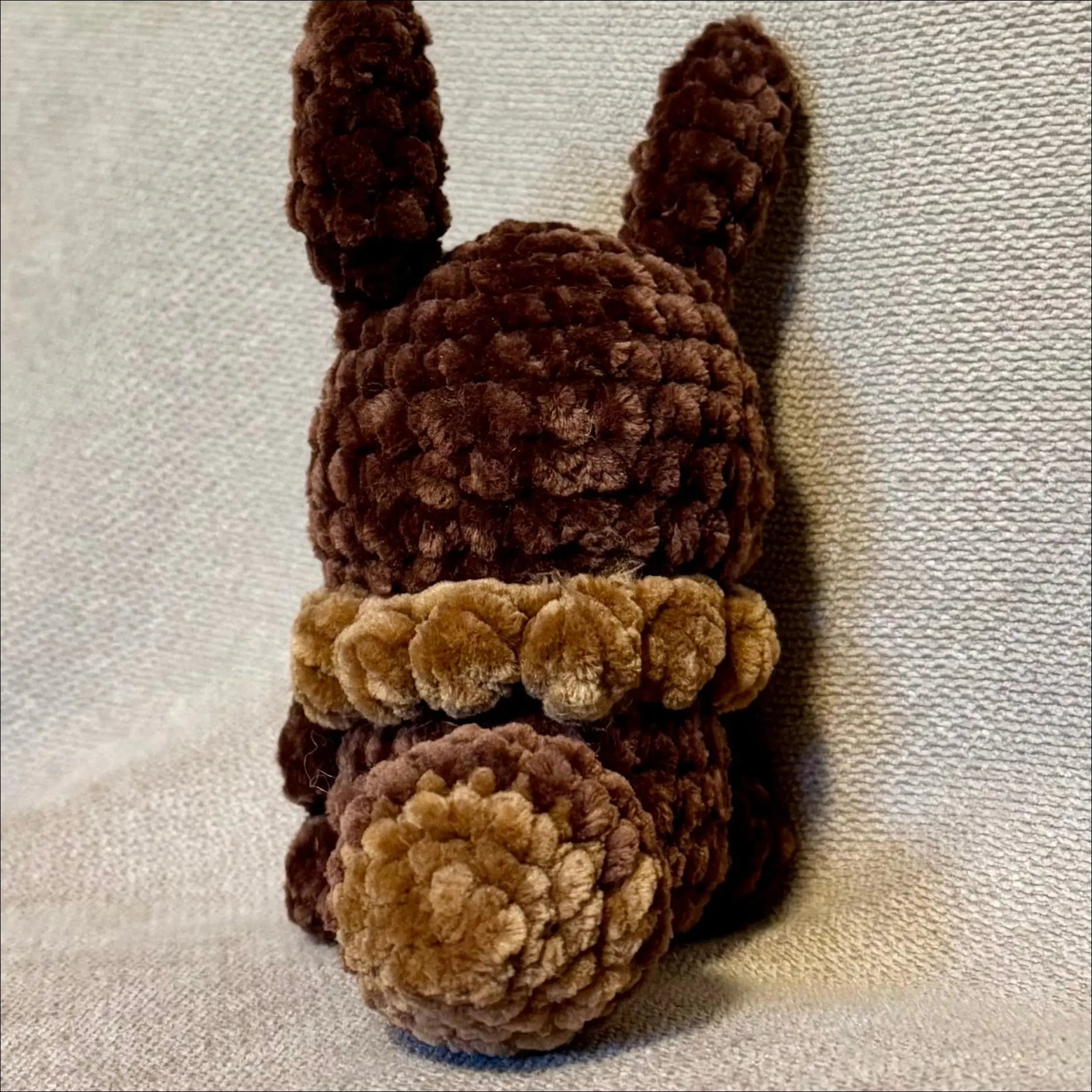 A small crocheted Eevee plush toy shown from the back. The toy is made from dark brown and light tan bouclé-like yarn, with upright ears, a dark brown body, and a light tan band around the middle.