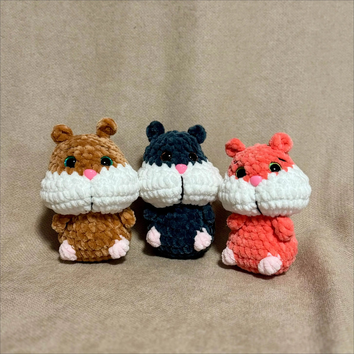 Three small crocheted hamster toys in brown, navy blue, and coral red, arranged side by side on a beige fabric background.