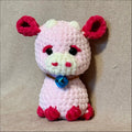 A crocheted plush toy shaped like a cute cow with a light pink body, dark pink ears and feet, a cream-colored snout and inner ears, black bead eyes, and a shiny blue bell around its neck, set against a neutral beige textured fabric background.