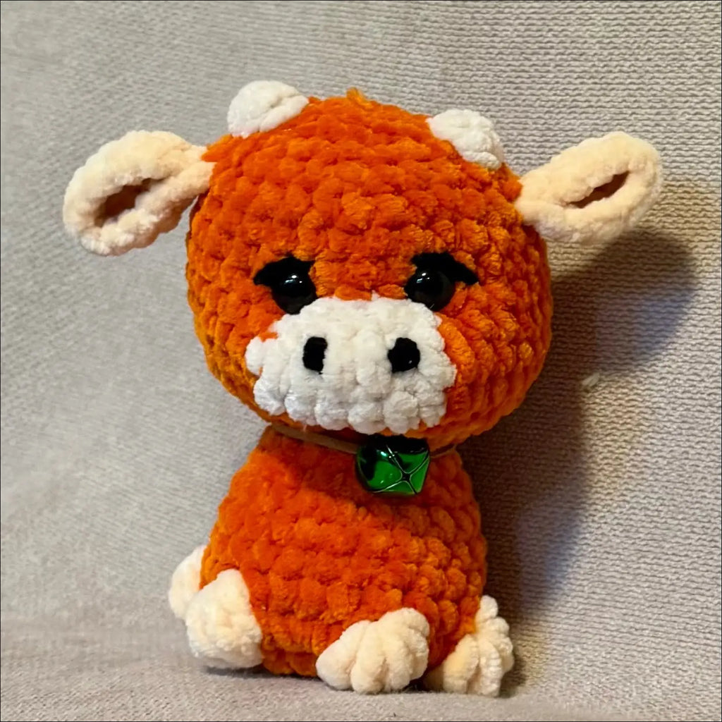 A small crocheted plush toy shaped like a cow, made from orange yarn with cream-colored ears, muzzle, and feet, and a small green bell around its neck, sitting against a textured gray fabric background.