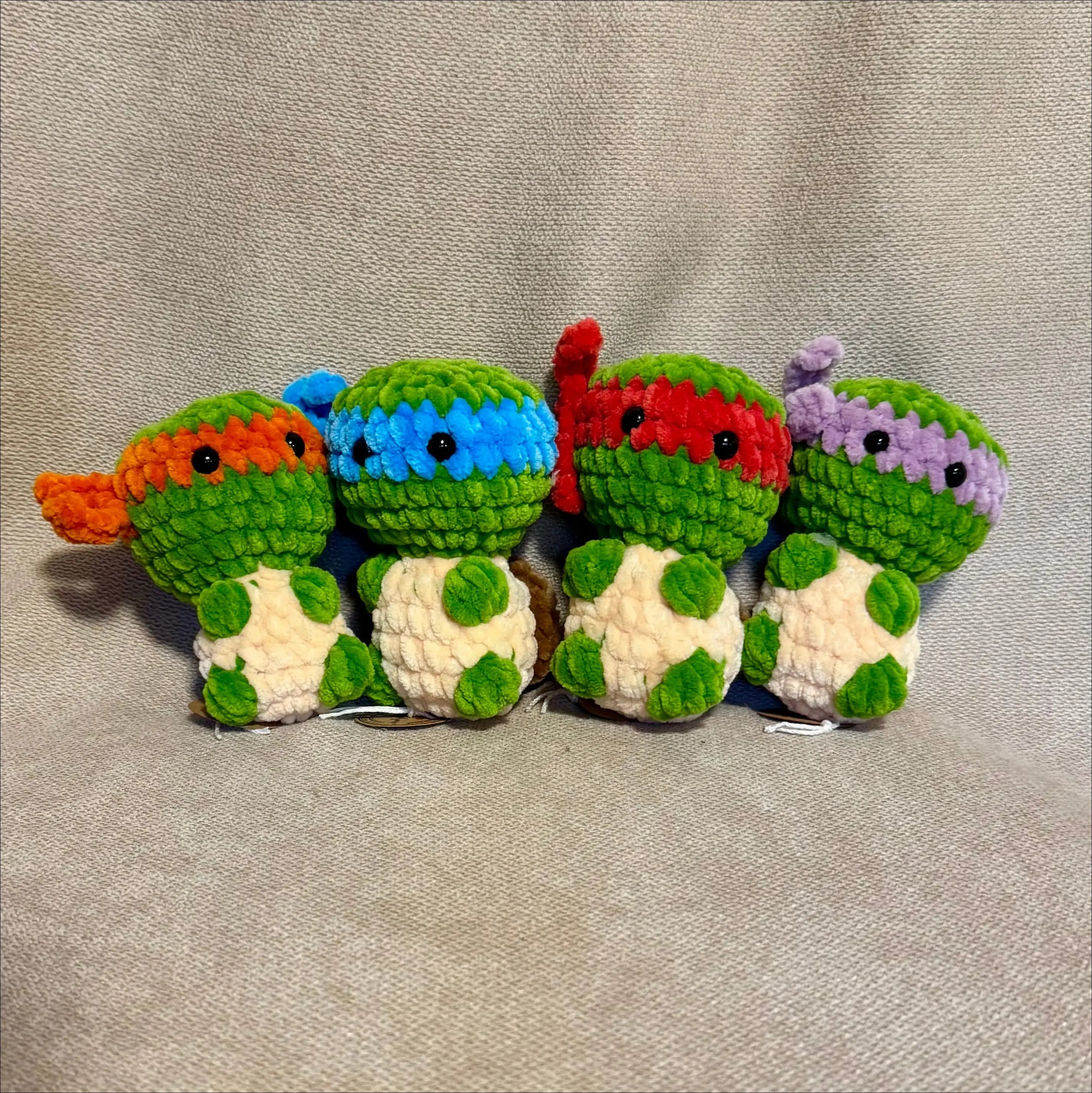 Four small crocheted turtle characters lined up on a light beige fabric background. Each turtle has a different colored head mask (orange, blue, red, and purple) and a white body with green shell patterns.