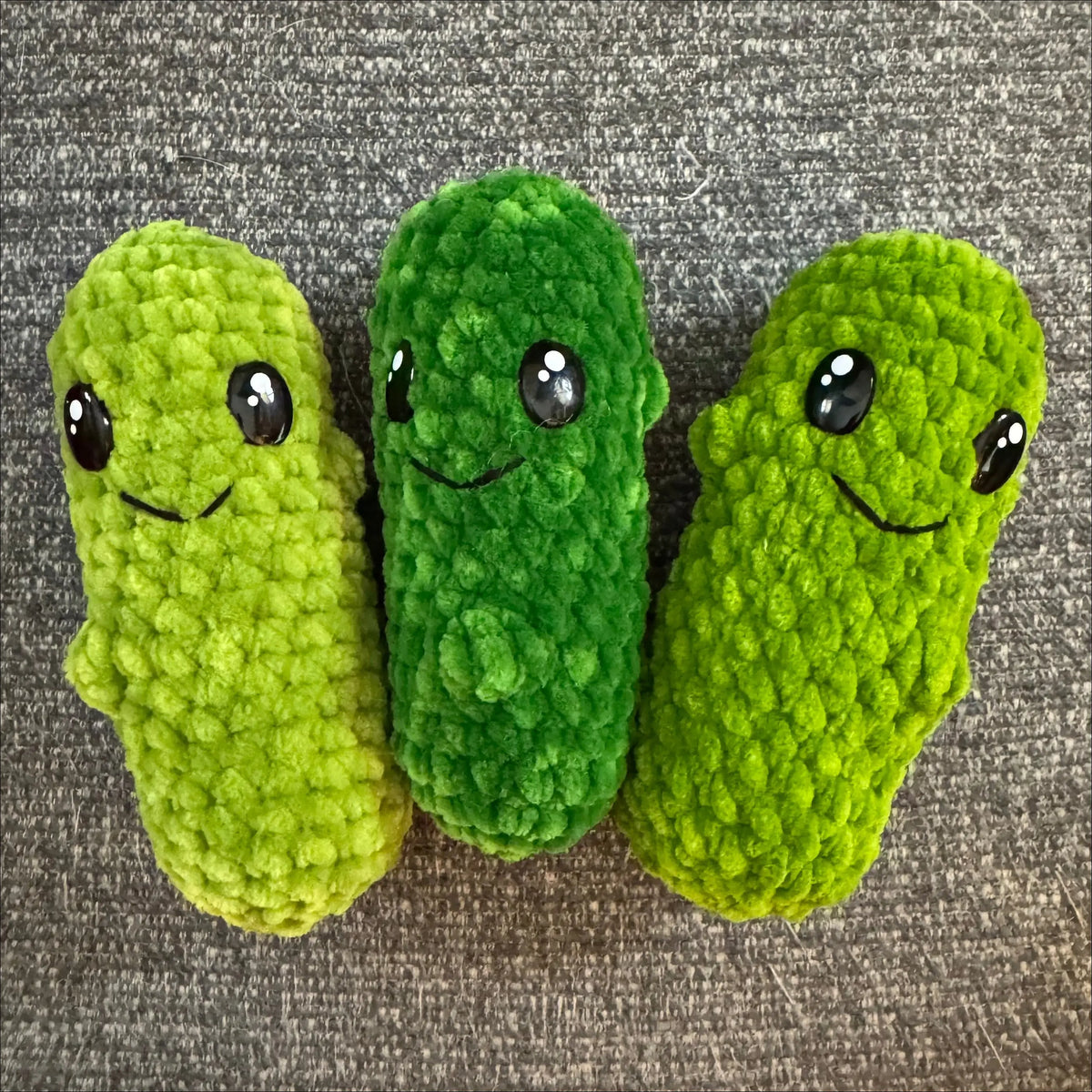 Three small crocheted pickle toys in different shades of green, each with embroidered eyes and a simple smile, arranged on a gray textured fabric background.