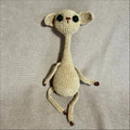 A handmade crocheted cat with large black eyes, cream-colored yarn, and small brown feet, lying on a textured beige fabric background.