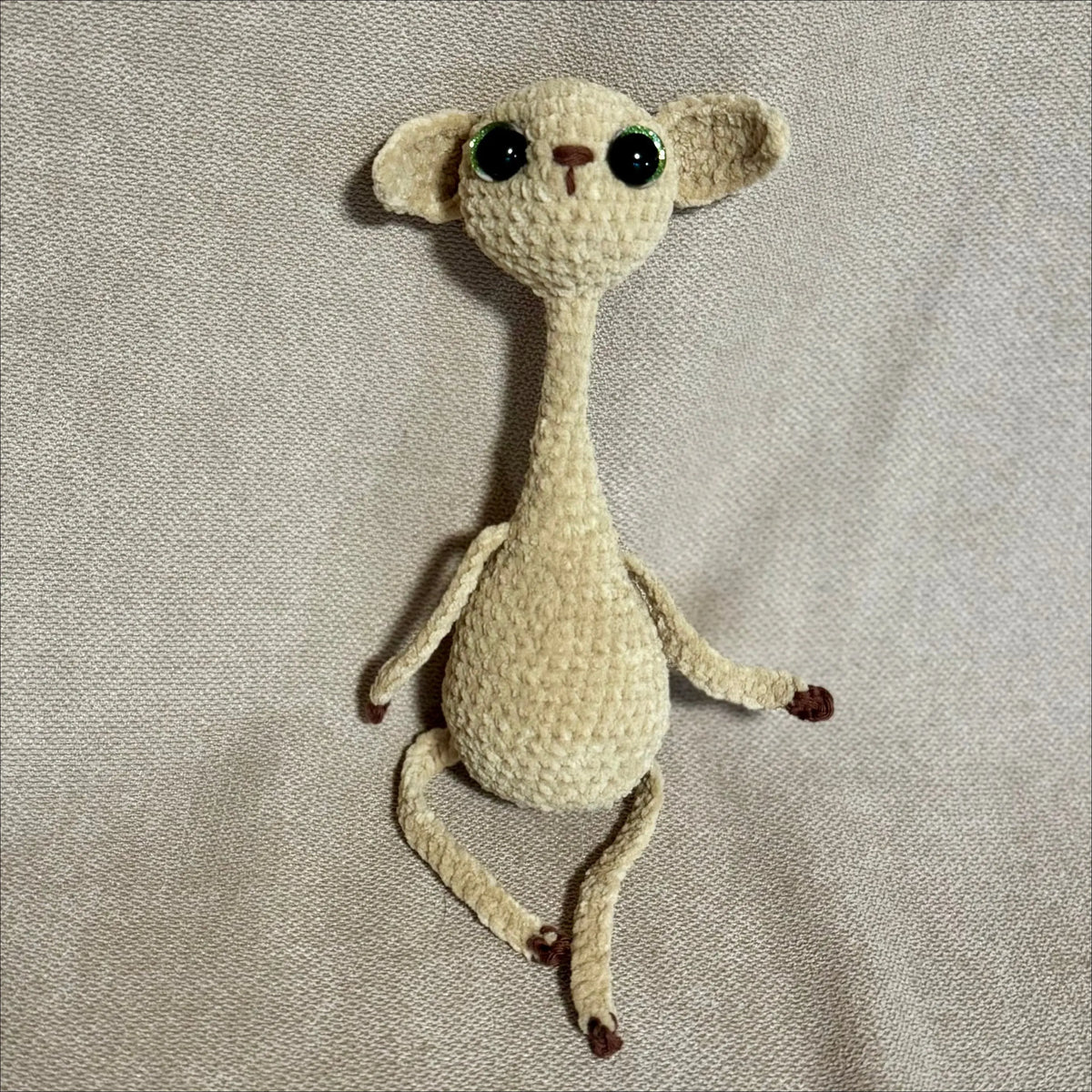 A handmade crocheted cat with large black eyes, cream-colored yarn, and small brown feet, lying on a textured beige fabric background.