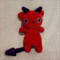 A small crocheted devil plush toy in bright red with dark purple horns and tail, featuring large black-and-white bead eyes, photographed on a beige fabric background.