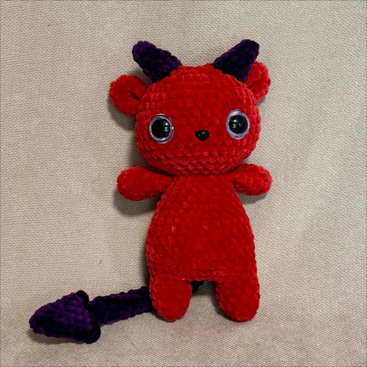 A small crocheted devil plush toy in bright red with dark purple horns and tail, featuring large black-and-white bead eyes, photographed on a beige fabric background.