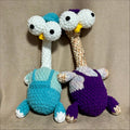 Two handmade crocheted toy chicken with long necks, displayed against a neutral beige fabric background. One toy is light blue and white, and the other is purple and beige.