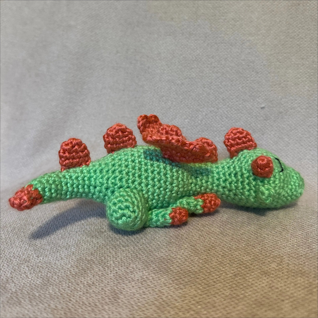 A small crocheted dragon  toy in light green with bright orange-red spikes and feet, resting on a textured beige fabric background.
