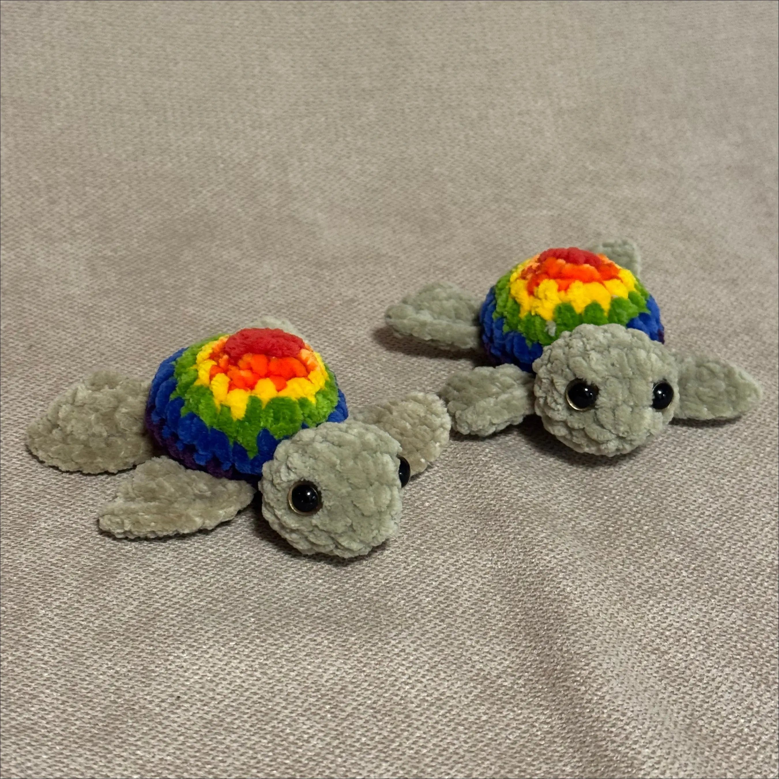 Two plush toys shaped like turtles with rainbow shells on a beige fabric background