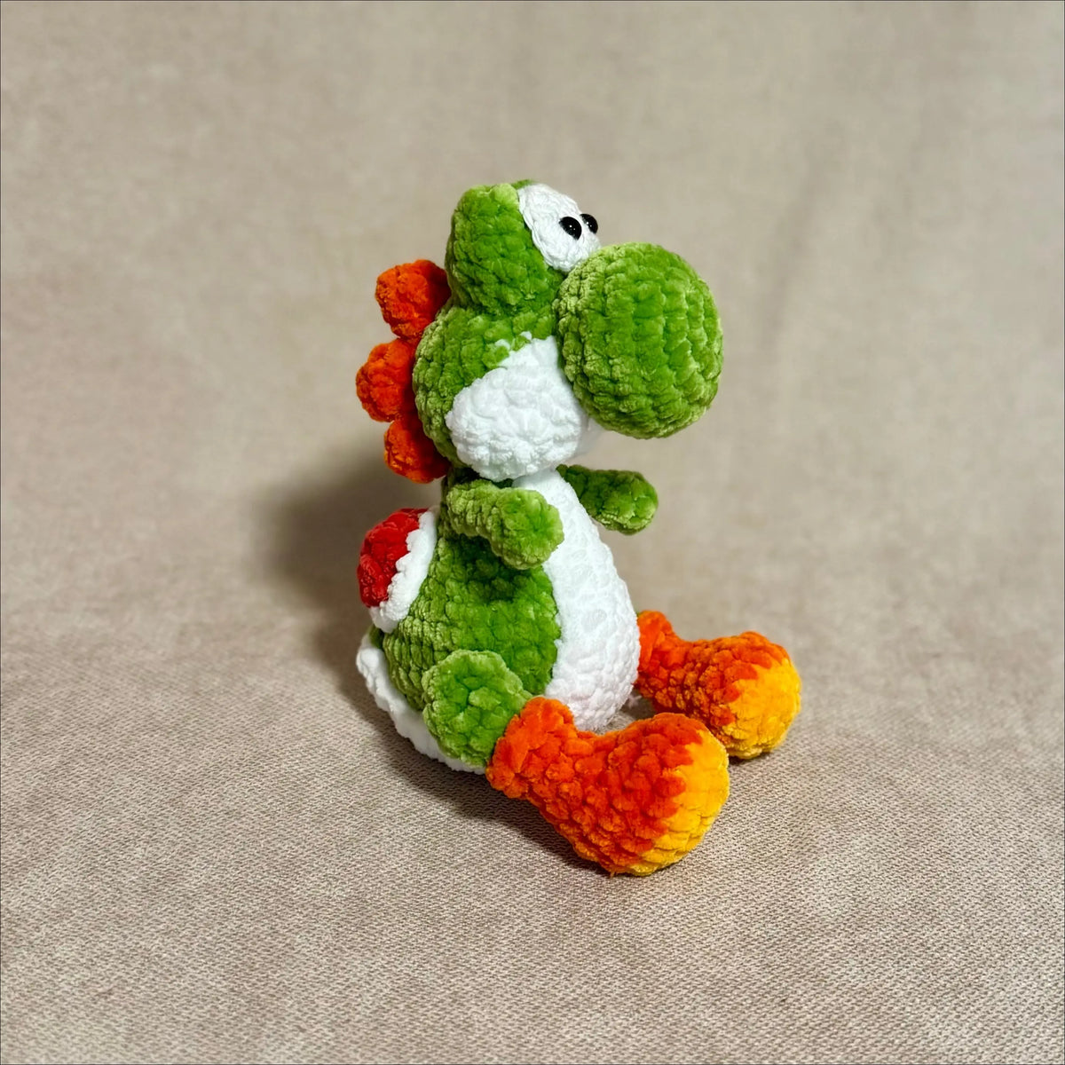A handmade crocheted toy resembling Yoshi, featuring green, white, orange, and red colors, sitting on a beige textured fabric background.